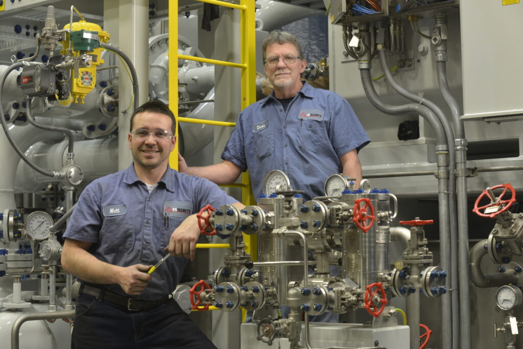 Two FS-Elliott technicians wearing safety glasses and blue work uniforms pose beside industrial compressor equipment with stainless steel piping, pressure gauges, and valves.