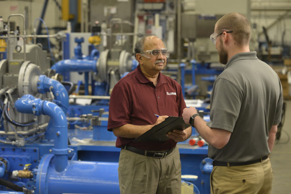 Two FS-Elliott employees wearing safety glasses discuss technical details while reviewing a clipboard in an industrial compressor manufacturing facility with blue machinery in the background.